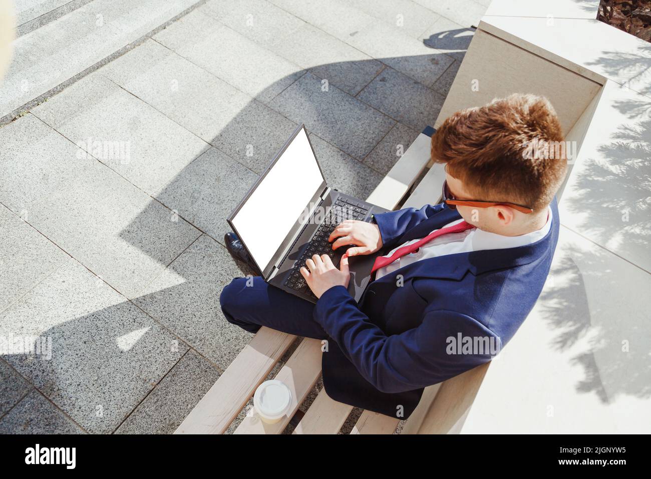 Junger Mann im Anzug mit Laptop mit leerem Bildschirm sitzt auf Steinbank. Blick von oben, Mockup. Mann mit Laptop für Online-Arbeit, Börse tr Stockfoto