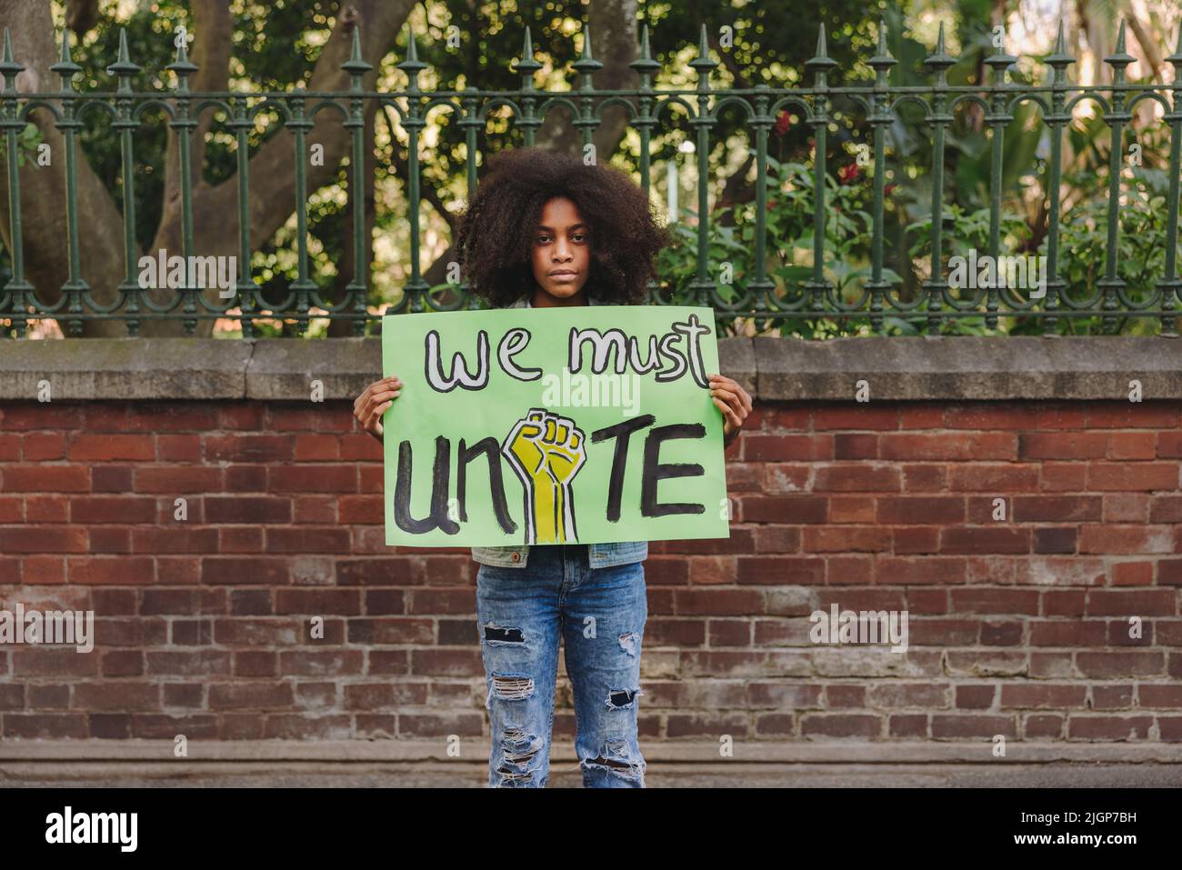 Schwarzes Teenager-Mädchen, das die Kamera anschaut, während es ein Plakat hält, das zur Einheit aufruft. Jugendaktivistin protestiert gegen Rassenungleichheit und kritisiert Stockfoto