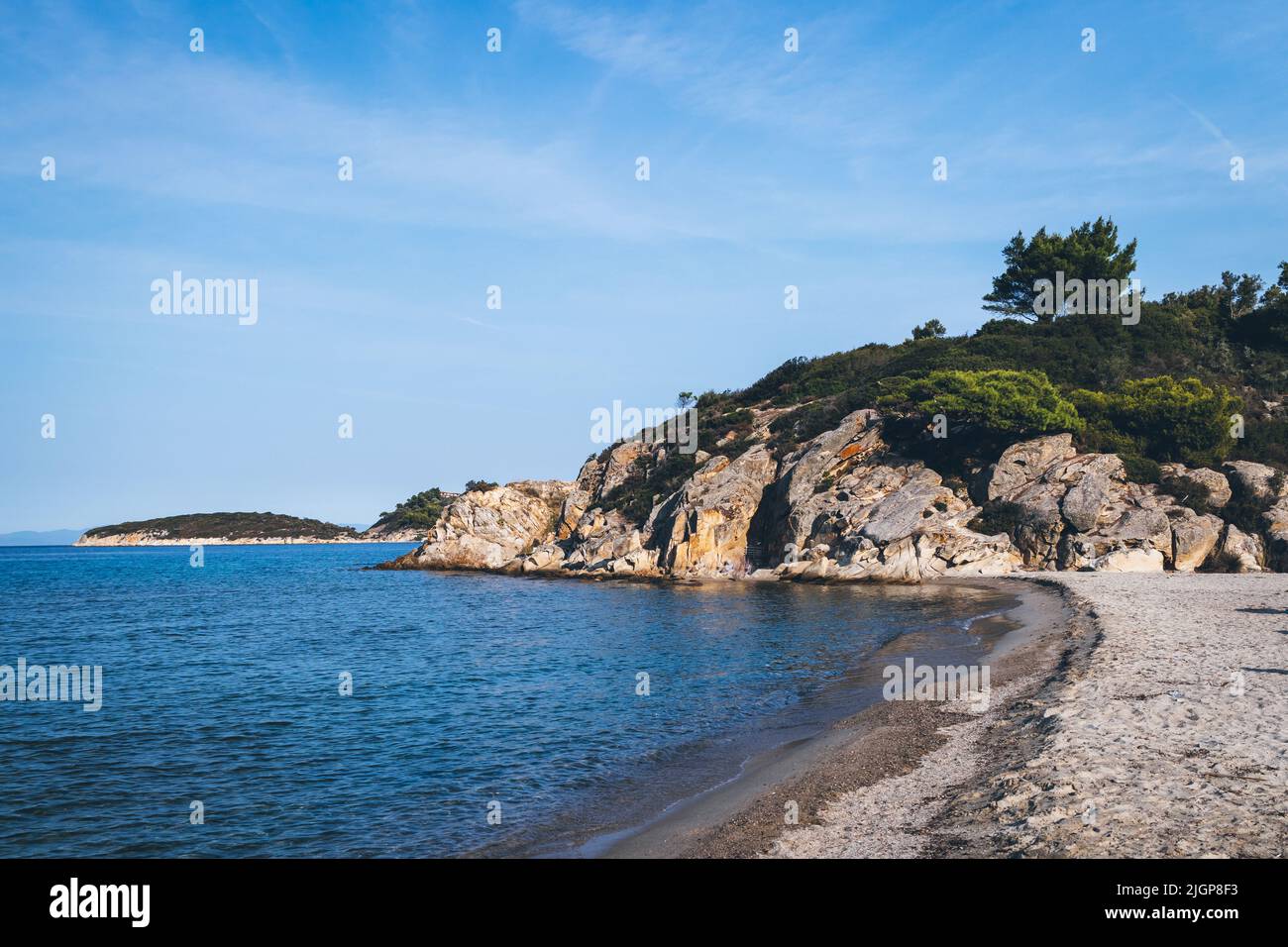 Blick auf den Küstenstrand im Hintergrund ein Felsvorsprung, der mit grünen Bäumen und Sträuchern bedeckt ist. Ormos Panagias, Griechenland. Stockfoto