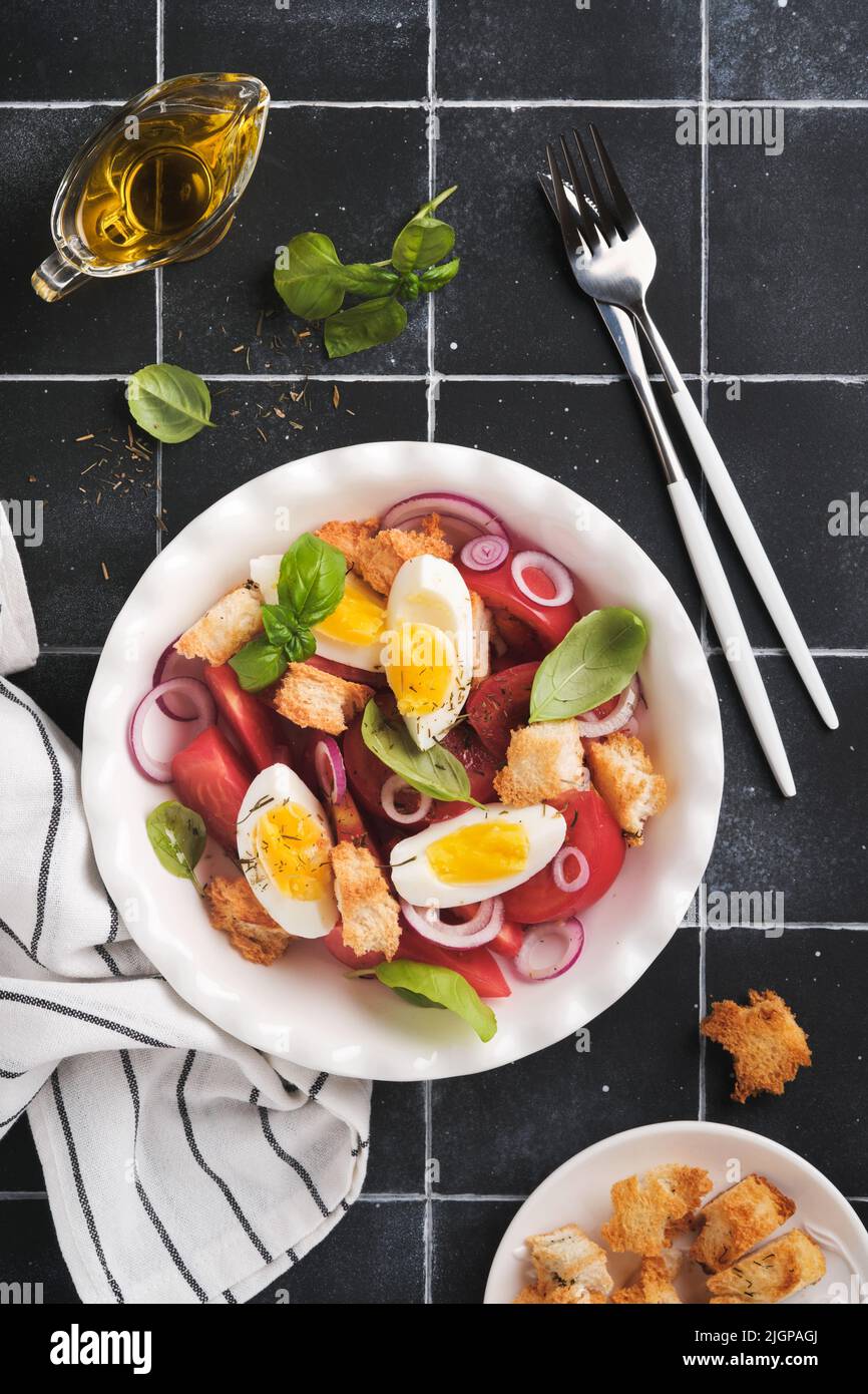 Toskanischer Panzanella-Salat. Frischer Tomaten-Salat mit Basilikumblättern, Eiern, Olivenöl und Zwiebeln in einer weißen Schüssel auf altem schwarzen Hintergrund. Vegetarisches Panzanell Stockfoto