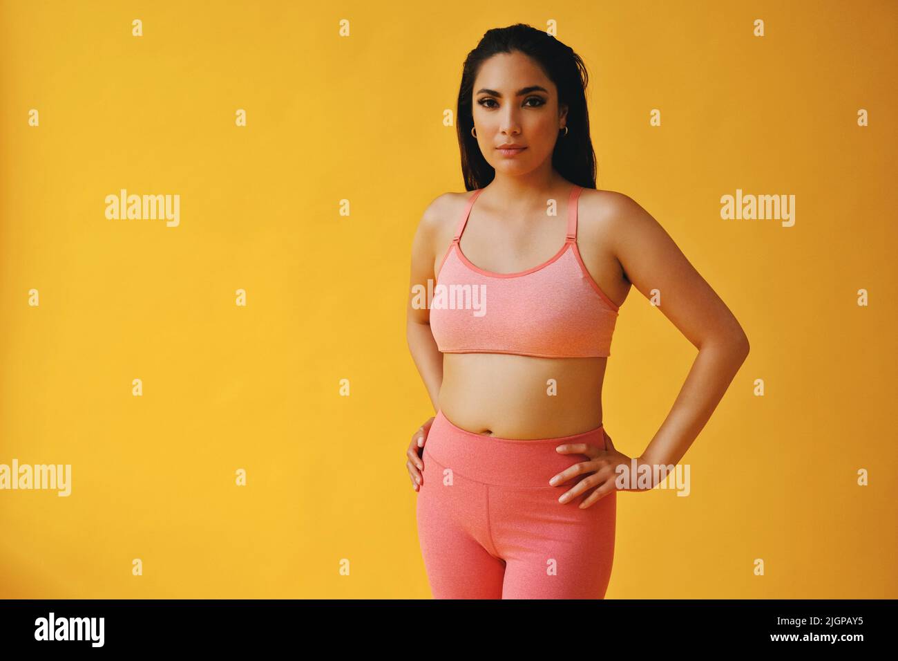 Waist-up-Porträt der hispanischen Schönheit Latina Frau mit Händen auf Hüften Sport-Outfit jungen Erwachsenen vor gelbem Hintergrund Blick auf Kamera-Studio aufgenommen Stockfoto