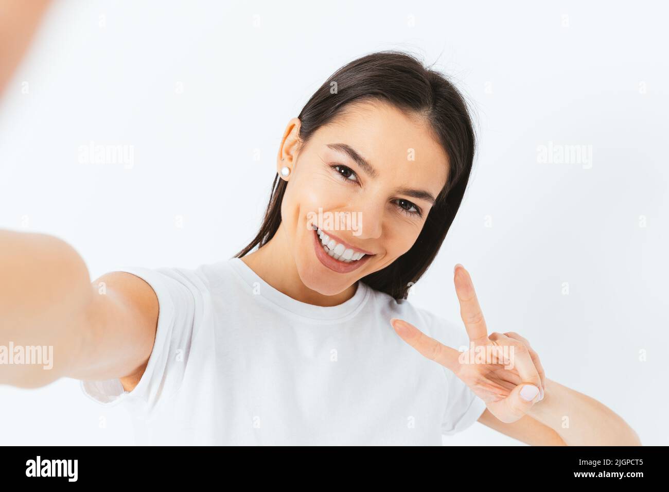 Porträt einer glücklichen jungen Frau macht Selfie zeigt zwei Finger Sieg Geste zur Kamera, weißer Hintergrund Stockfoto
