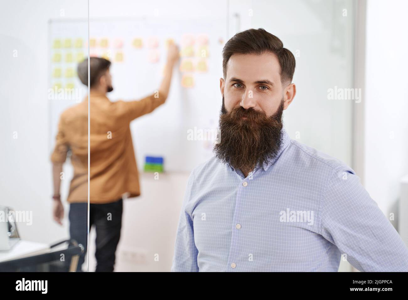 Glückliches Lächeln bärtige Senior Developer oder Manager in modernen IT-Büro. Ein weiterer Profi, der mit Scrum Desk im Hintergrund arbeitet. Erfolgreich Stockfoto