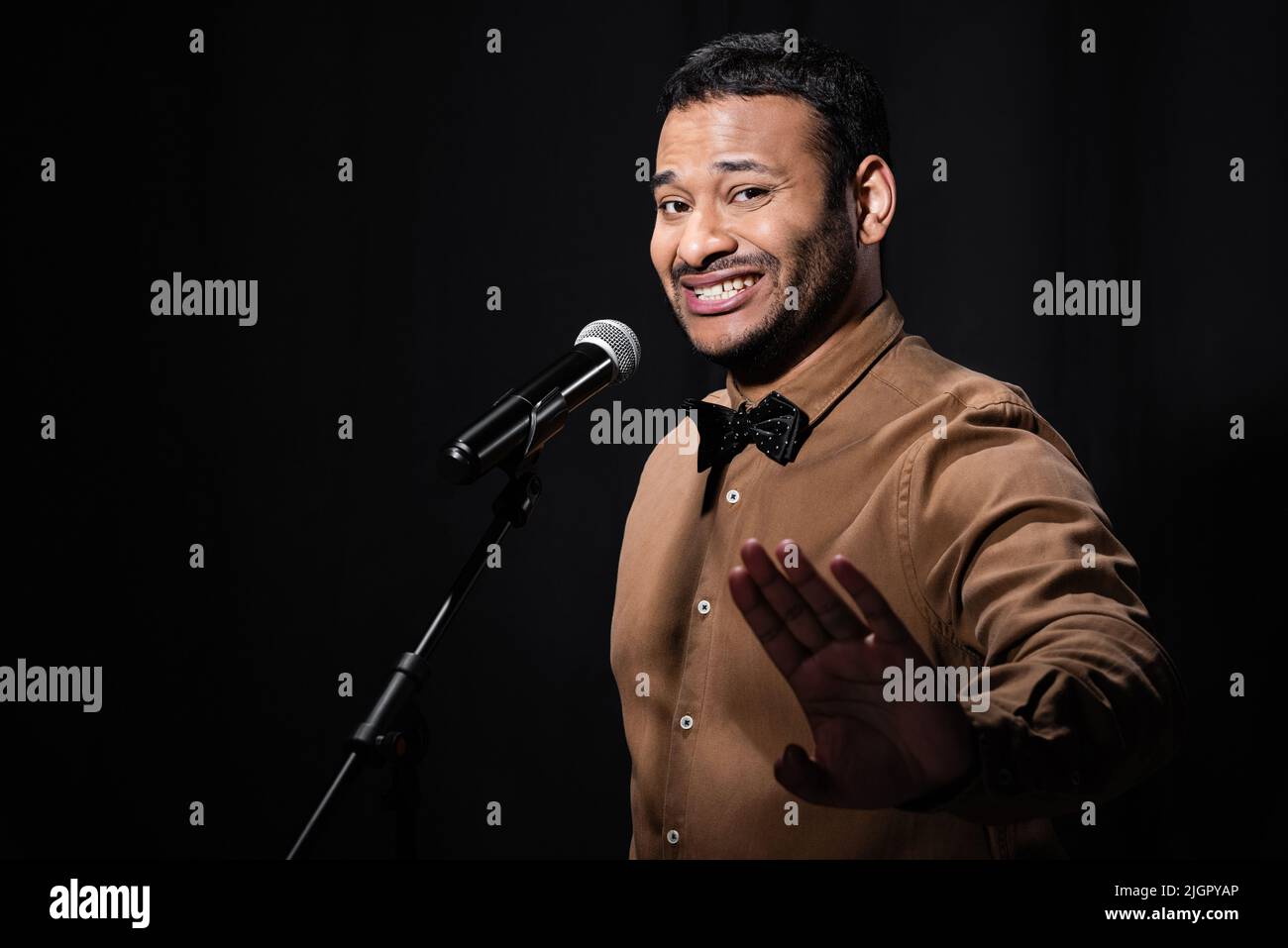 Unzufriedener indischer Stand-up-Komiker, der eine Stop-Geste zeigt und dabei Witze in ein schwarz isoliertes Mikrofon erzählt Stockfoto
