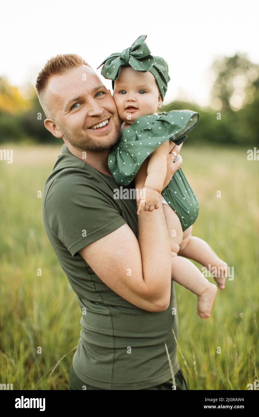 Porträt eines jungen Mannes, der lächelt und die Kamera anschaut, während er während des Picknicks auf der Wiese mit seiner Tochter auf den Händen spazierengeht. Vater genießt jeden Moment mit seinem liebenden Kind. Stockfoto