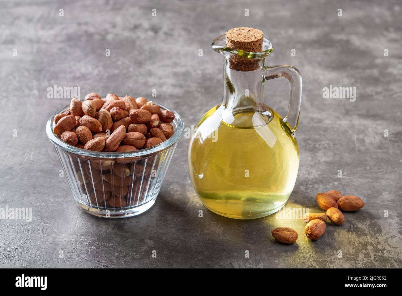 Erdnussöl in einem Glaskrug und rohe geschälte Erdnuss in einer Glasschale auf grauem Hintergrund. Arachis hypogaea als essbare Samen und Ölpflanzen. Nahaufnahme. Stockfoto