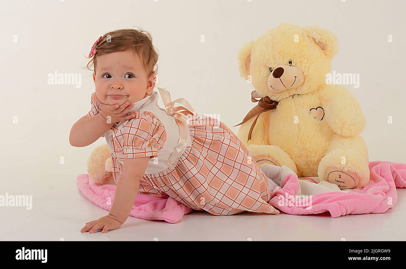 Ein kleines Mädchen in einem rosa karierten Kleid kriecht von einem Bären ein gelber Teddybär sitzt auf einem weißen Hintergrund das Mädchen beißt ihre Faust und schaut in den Rahmen. Hochwertige Fotos Stockfoto