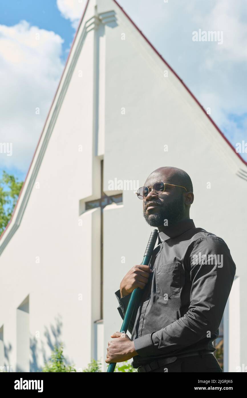 Junger ernsthafter Pastor der lutherischen Kirche in Brillen und schwarzem Hemd mit Bürokragenkragen, der Rechen gegen weißes Gebäude mit Kreuz hält Stockfoto