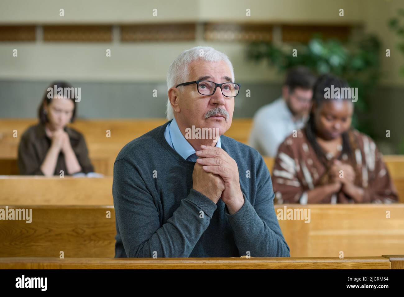 Gealterter Mann in Casualwear und Brillen, der seine Hände während des stillen Gebetes zusammenhält, während er das Kreuz in der Kirche anschaut Stockfoto