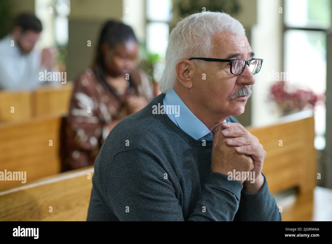 Reifer, männlicher Gemeindemitglied mit grauem Haar, der mit zusammengerissene Hände betet, während er auf der Bank gegen andere Menschen sitzt Stockfoto