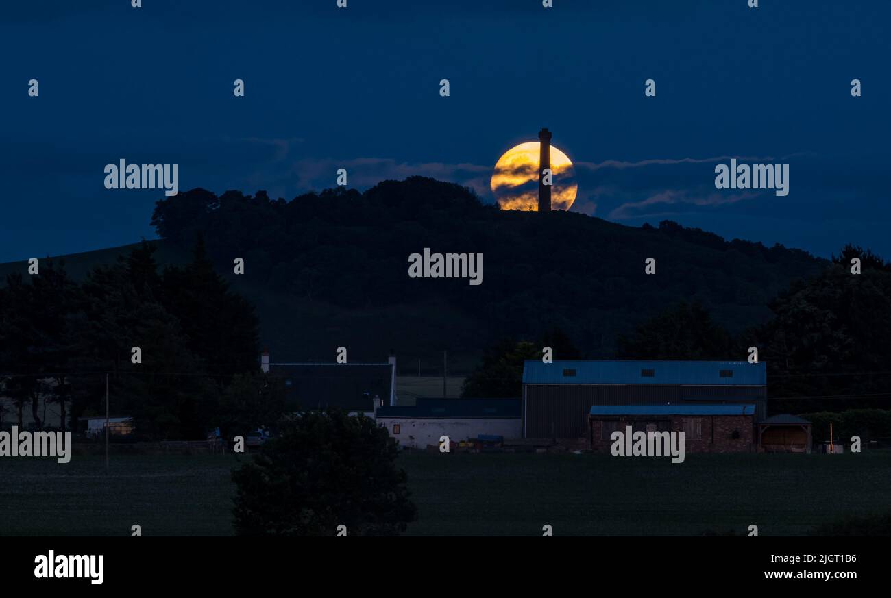 East Lothian, Schottland, Vereinigtes Königreich, 12.. Juli 2022. UK Wetter: Buck-Supermond: Der letzte Supermond in diesem Jahr ist der Vollmond im Juli, der Buck-Mond genannt wird, der neben dem viktorianischen Denkmal auf dem Hügel auf dem Byres Hill zu sehen ist. Der Mond liegt bei 98,7% seiner maximalen Größe Stockfoto