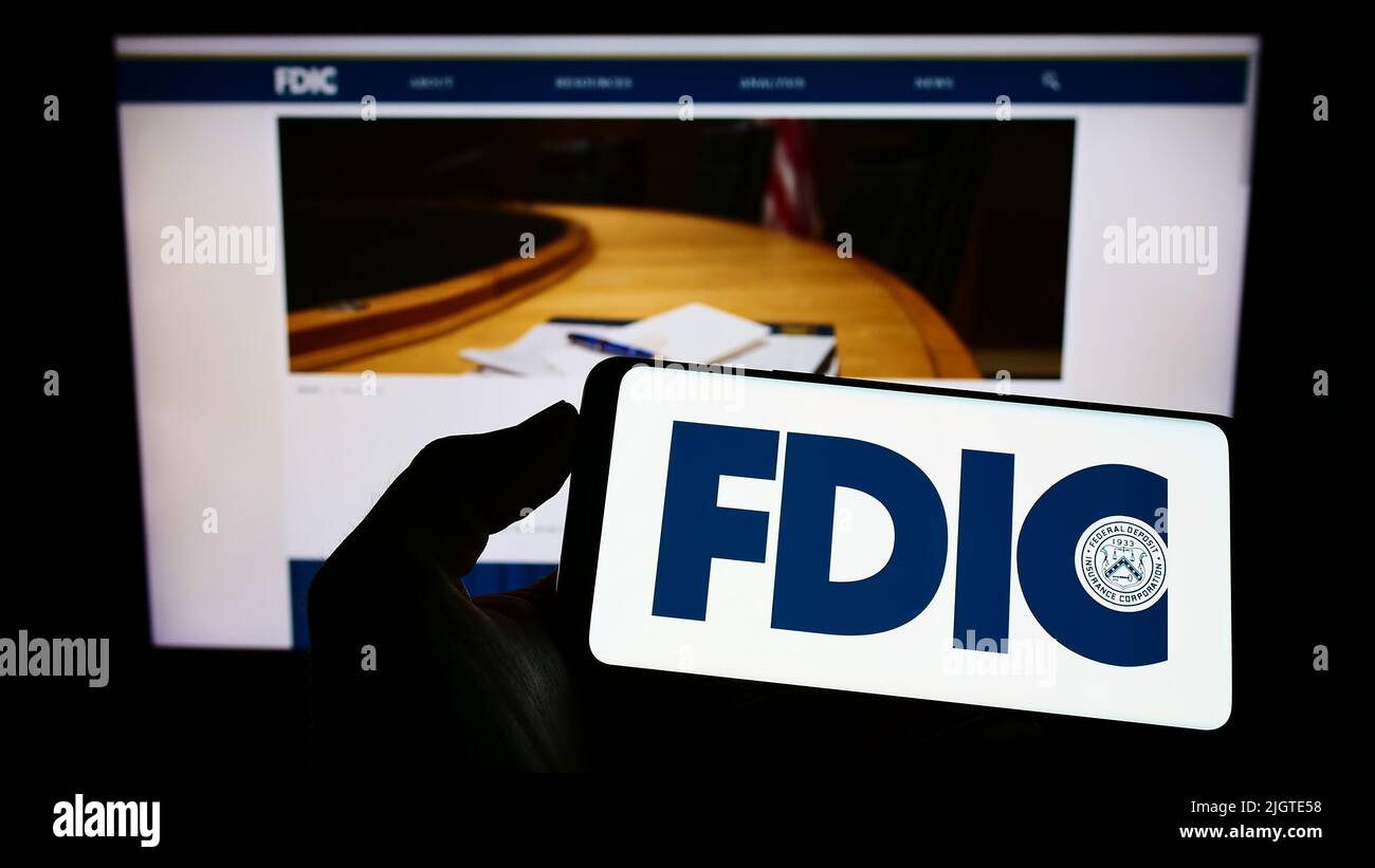 Person mit Mobiltelefon und Logo der US Federal Deposit Insurance Corporation (FDIC) auf dem Bildschirm vor der Webseite. Konzentrieren Sie sich auf die Telefonanzeige. Stockfoto