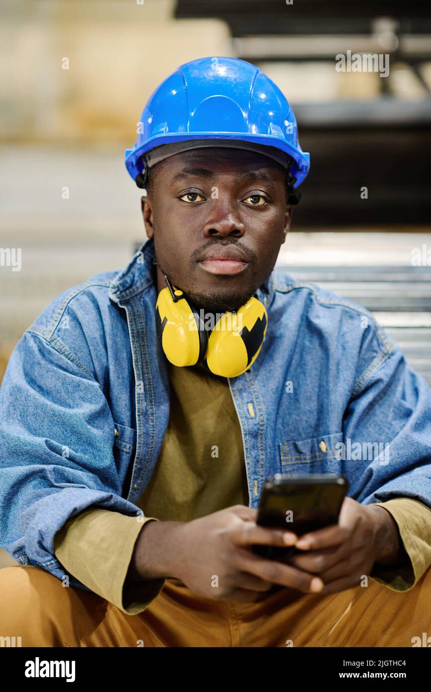 Porträt eines jungen afrikanischen Arbeiters in schützendem Hardhut, der während der Pause auf seinem Smartphone auf die Kamera schaut Stockfoto