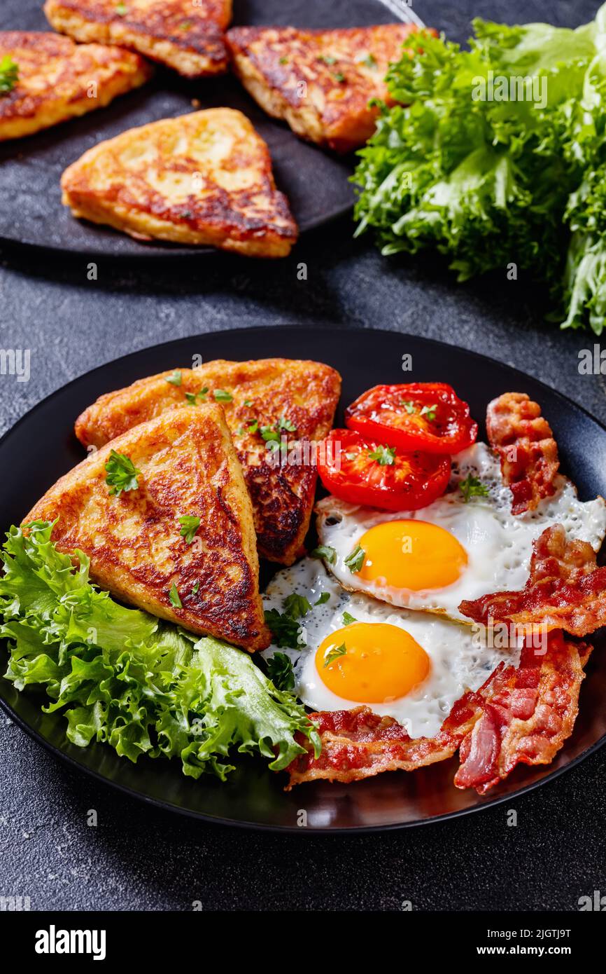 irisches Frühstück mit Spiegeleiern, Rascher- oder Speckscheiben, gebratenen Kartoffelkuchen, gegrillten Tomaten und frischem Salat auf schwarzem Teller auf Betontisch, vertic Stockfoto