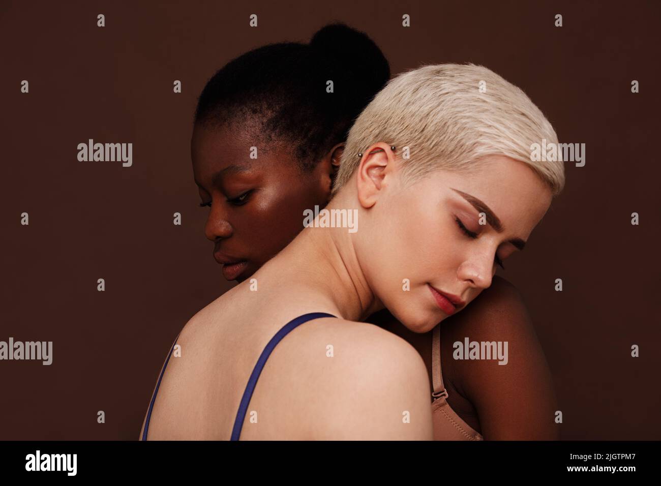 Zwei junge Frauen mit unterschiedlicher Hautfarbe stehen zusammen. Weibchen legen sich mit geschlossenen Augen den Kopf auf die Schultern. Stockfoto