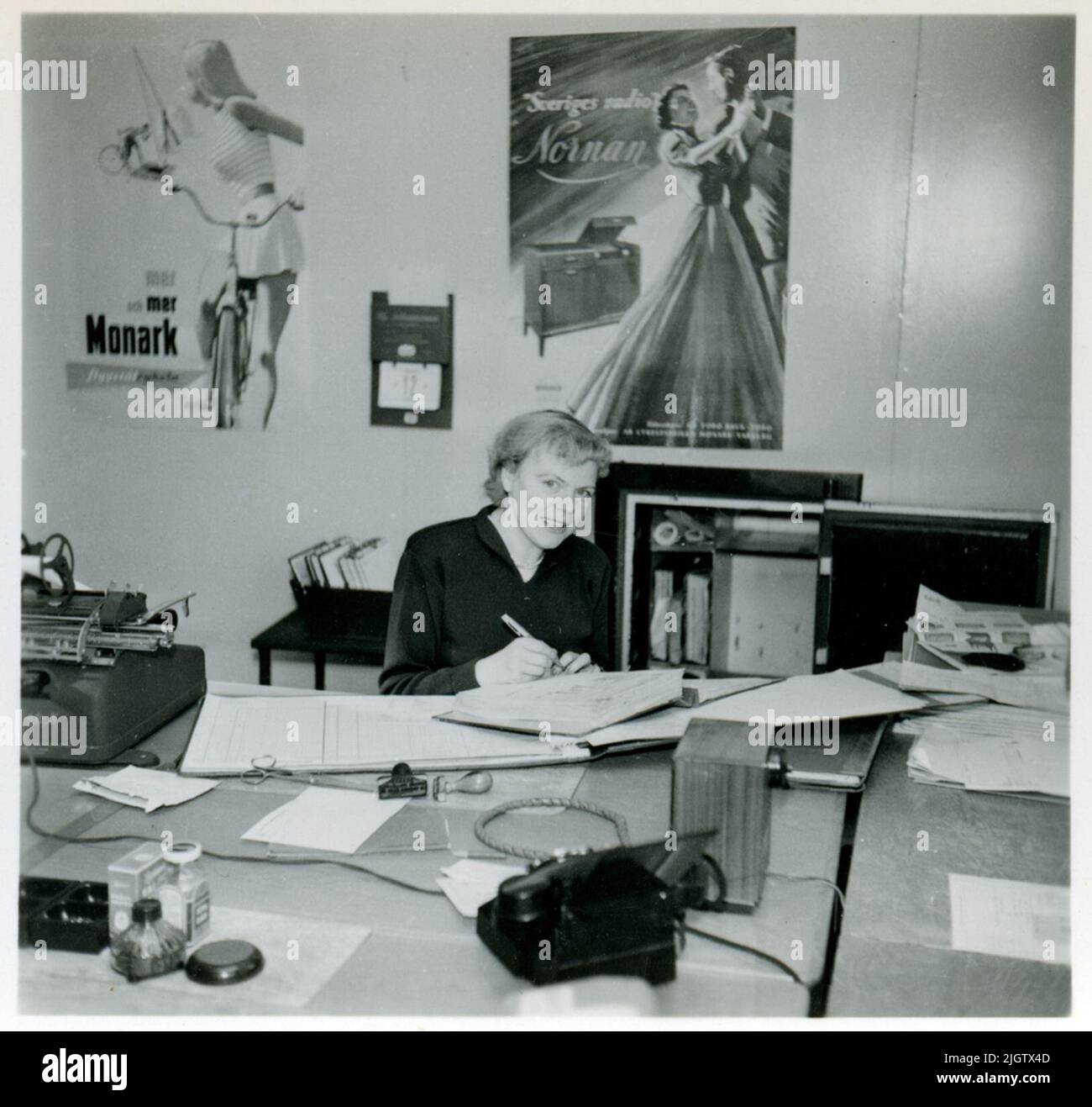 Das Büro in der Fahrradwerkstatt von Monarch. Eine Frau sitzt an einem Schreibtisch und schreibt in einem Cover. An der Wand hinter ihr befindet sich ein Plakat mit Werbung für Monarks Fahrräder und eines für Monarks Radiomöbel "Swedish Radio Nornan". Stockfoto