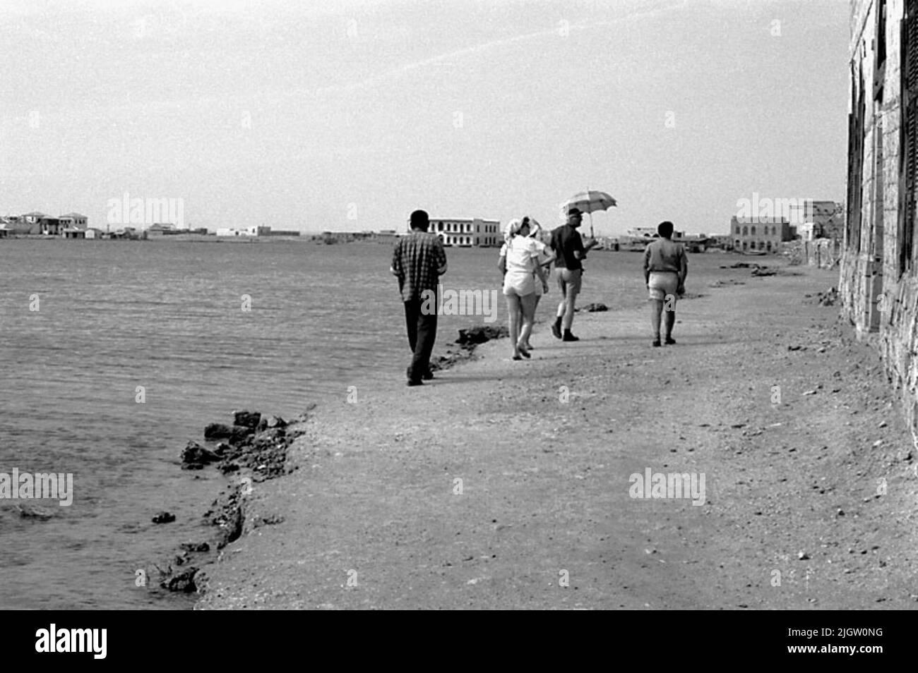 Das Foto wurde ungefähr 1961-08-22 aufgenommen. Drei Männer und zwei Frauen gehen an einem Strand spazieren und schauen auf eine teilweise ruinierte Gesellschaft. African travel35 Bilder in Serie. Stockfoto