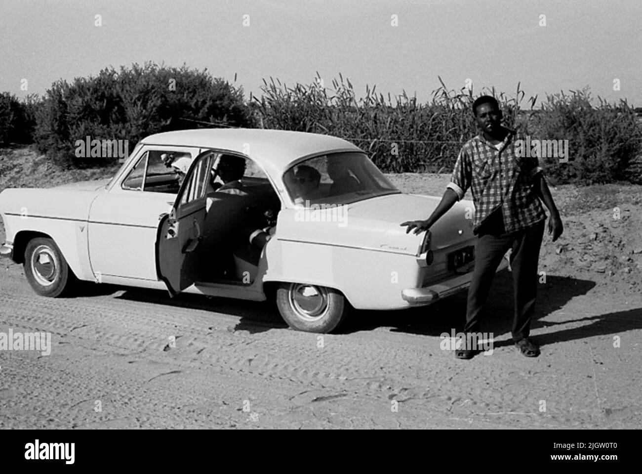 Das Foto wurde ungefähr 1961-08-22 aufgenommen. Ein Auto wird auf einer Straße geparkt. Rechts vom Auto steht ein Mann und lehnt sich zur Heckklappe. African travel35 Bilder in Serie. Stockfoto