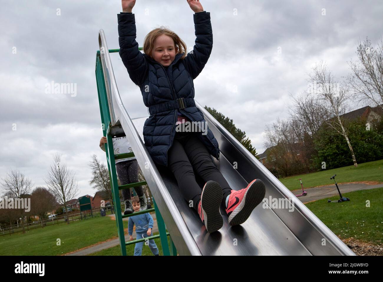 Ein junges Mädchen, das in Winterkleidung in einem Park in großbritannien auf einer Rutsche herunterrutscht und die Arme hochgezogen hat Stockfoto