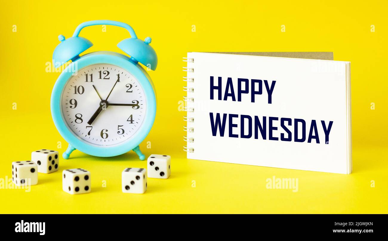 Auf gelbem Hintergrund ein blauer Wecker, Würfel und ein Notizbuch mit der Aufschrift HAPPY WEDNESDAY. Stockfoto