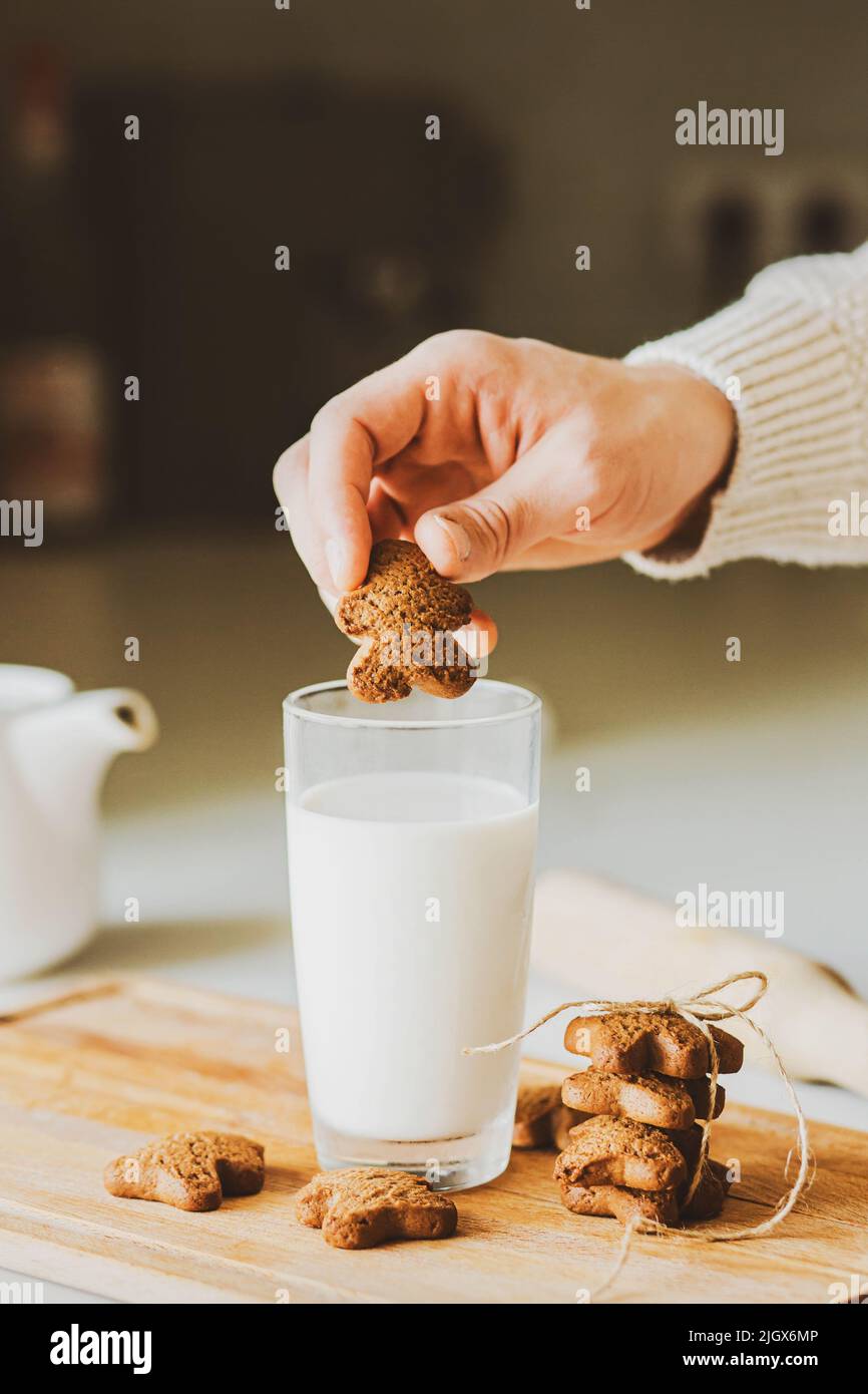 Ein Mann wird einen leckeren Haferflocken-Cookie in Form eines Lebkuchenmannes in ein Glas tauchen. Auf dem Küchentisch steht ein Glas Milch und Gin Stockfoto
