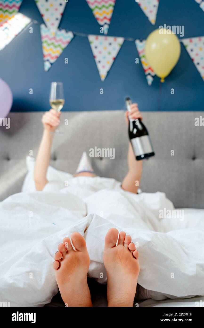 Selektiver Fokus auf die Füße einer Frau, die auf dem Bett liegt, mit erhobenen Händen, die Flasche und ein Glas Champagner in einem dekorierten Schlafzimmer halten. Pläne für Geburtstag. Zeit Stockfoto
