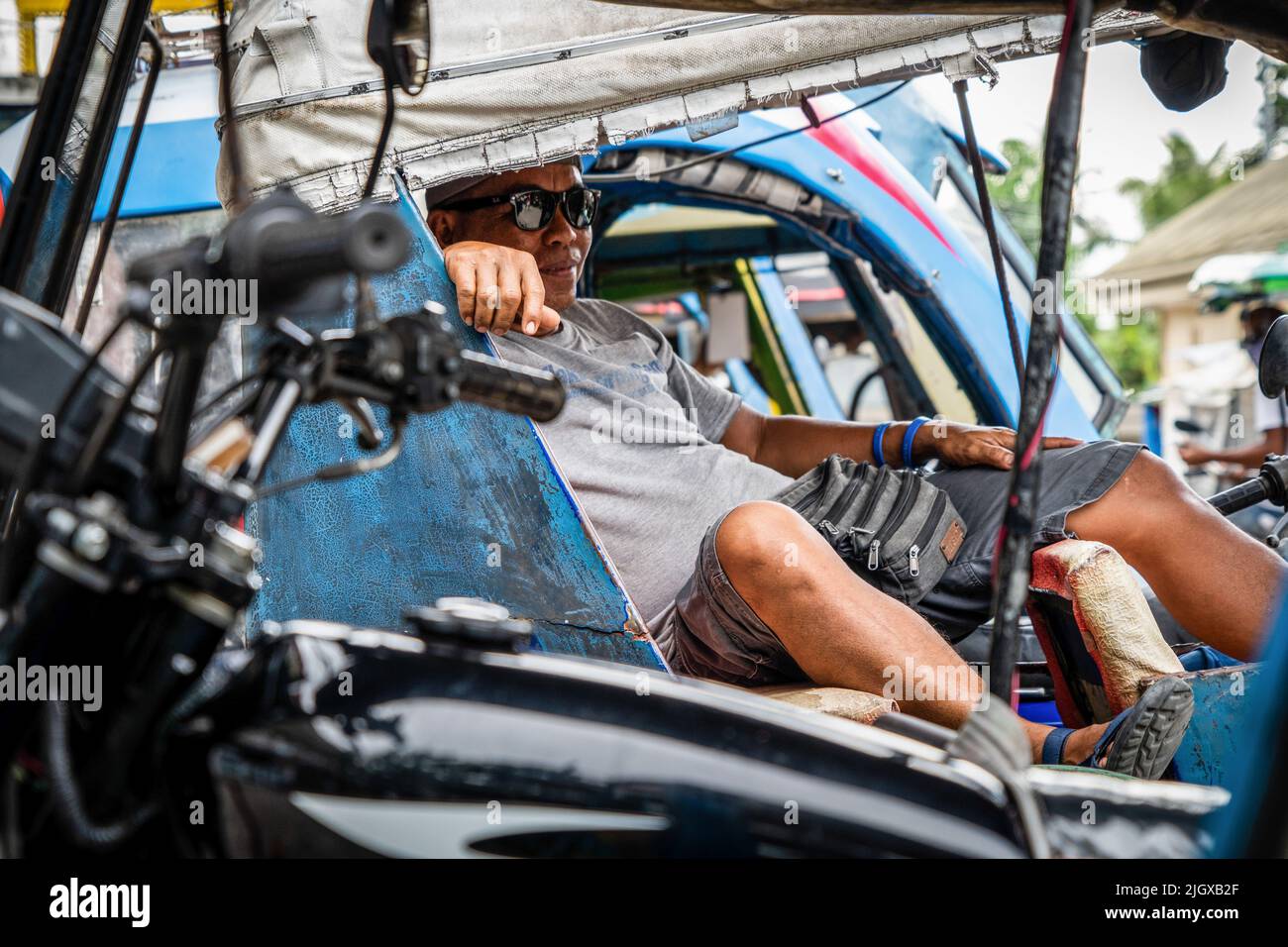 Moalboal, Philippinen. 13.. Juli 2022. Ein Motorrad-Dreirad-Fahrer macht eine Pause, während er vor einer belebten öffentlichen Bushaltestelle auf Kunden wartet. Tägliches Leben im Moalboal District, Cebu, Philippinen. Das Gebiet, das einst für sein Tauchen und seine massiven Sardinenwolken bekannt war, hat sich schwer getan, den Tourismus wieder zu beleben, auch nachdem die Reisebeschränkungen von Covid-19 aufgehoben wurden, da die Infrastruktur durch den Taifun Rai, auch bekannt als Super Typhoon Odette, beschädigt wurde, was einen Großteil des Gebiets dezimiert und mit dem Wiederaufbau zu kämpfen hat. Kredit: SOPA Images Limited/Alamy Live Nachrichten Stockfoto