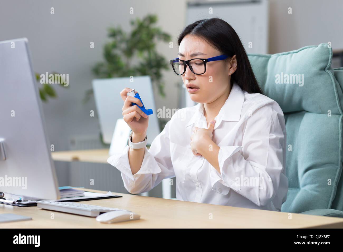 Allergie, Unwohlsein am Arbeitsplatz. Eine junge asiatische Frau in einer Brille hustet, würgt, nutzt einen Inhalator, niest in ihrem Hals, hat Allergien, Asthma. Er sitzt an seinem Schreibtisch im Büro. Stockfoto