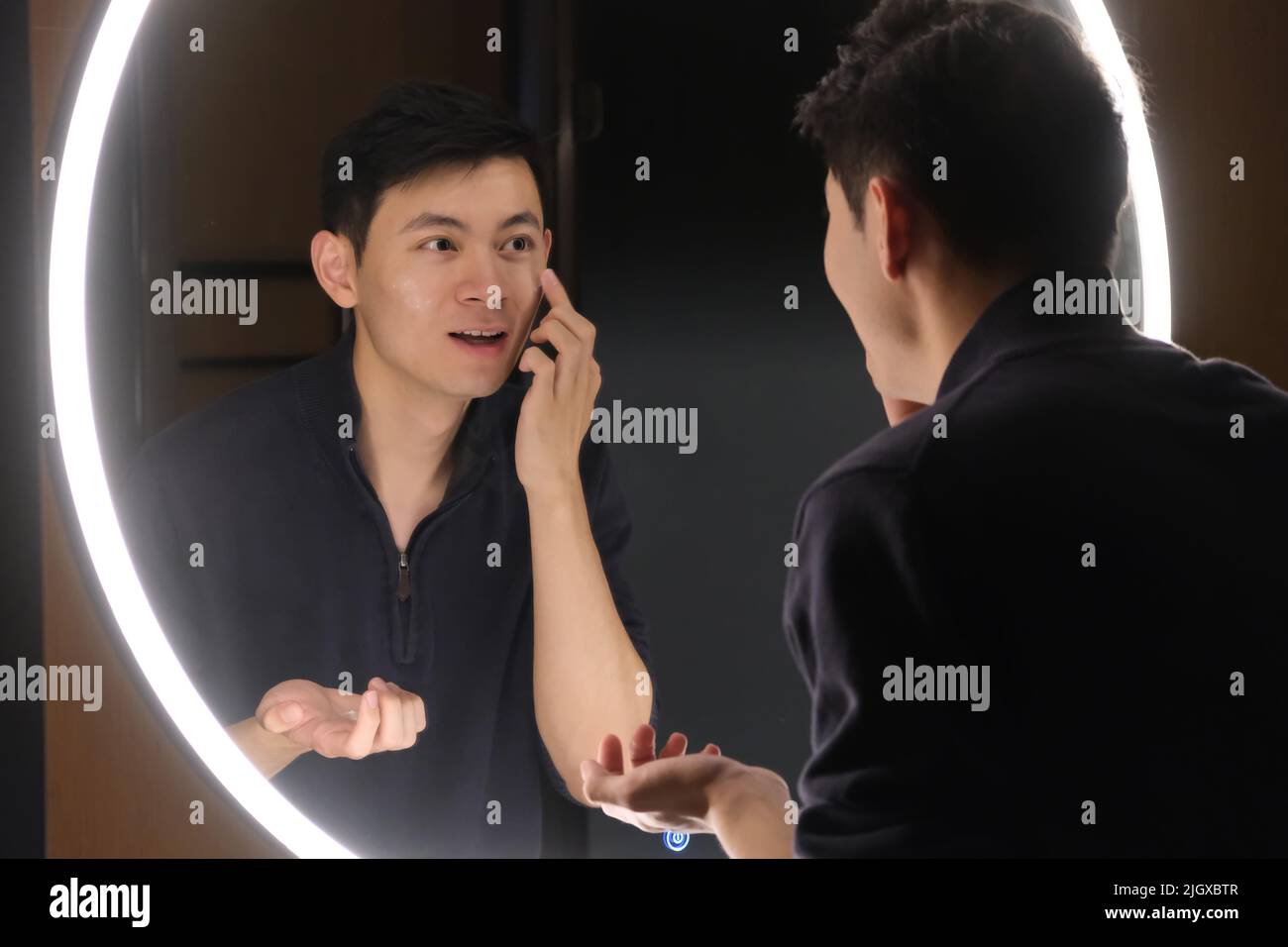 Ein hübscher junger asiatischer Mann trägt im Spiegel Hautpflegeprodukte auf das Gesicht auf Stockfoto