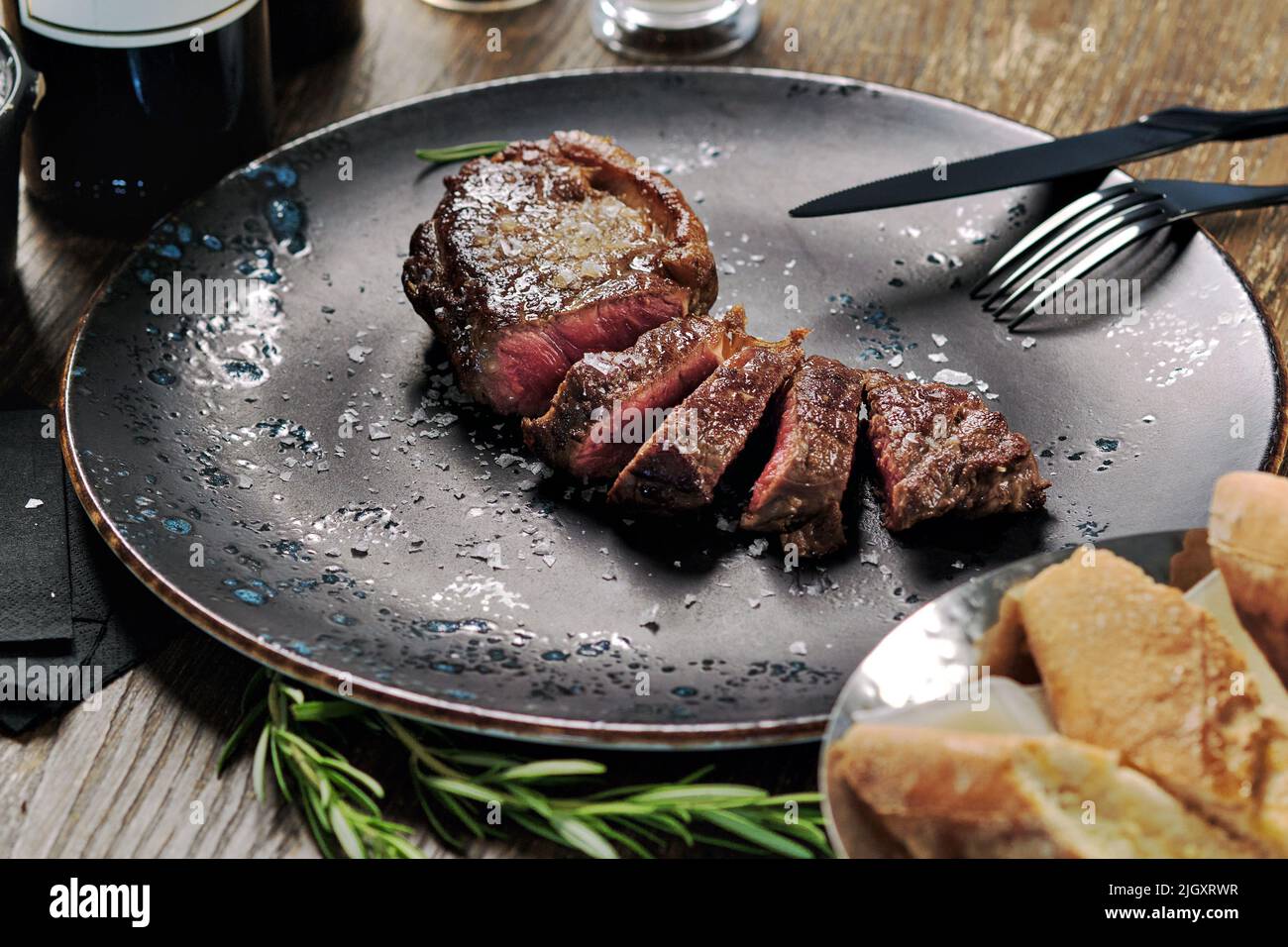 Nahaufnahme geschnittenes mittelseltenes Steak, serviert auf Teller mit Besteck und Schüssel mit frisch gebackenem Brot. Hauptgericht, Restaurantgericht, Esskonzept Stockfoto