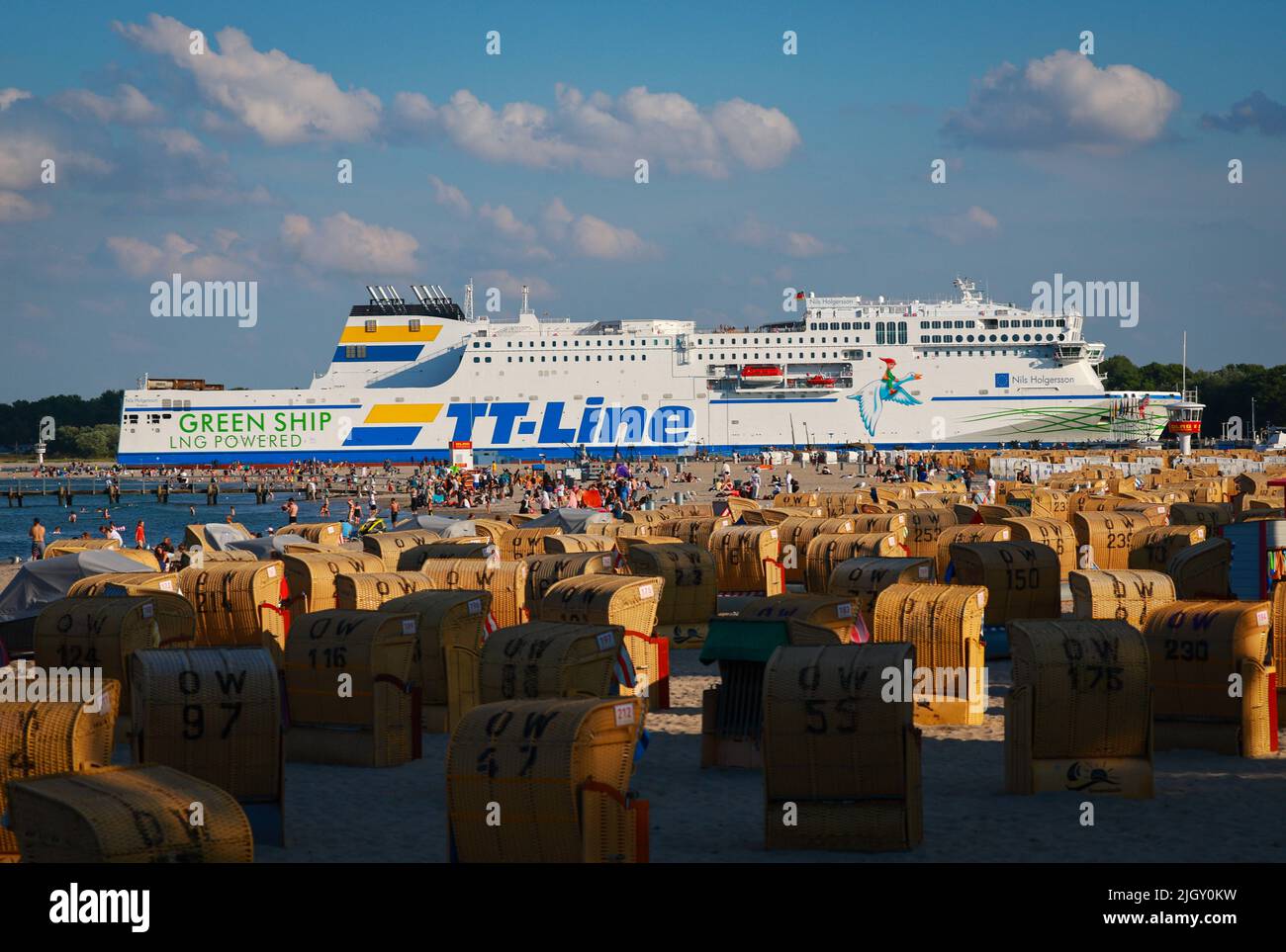 13. Juli 2022, Schleswig-Holstein, Travemünde: Hinter zahlreichen Liegestühlen voller Tagesausflügler und Urlauber fährt die Fähre Nils Holgerson von TT-Linie in den Hafen von Travemünde. Während große Teile Südeuropas unter Waldbränden, Dürre und extremer Hitze von bis zu 44 Grad leiden, können sich die Menschen im von Dürre heimgesuchten Deutschland auf hohe Sommertemperaturen freuen. Foto: Christian Charisius/dpa Stockfoto