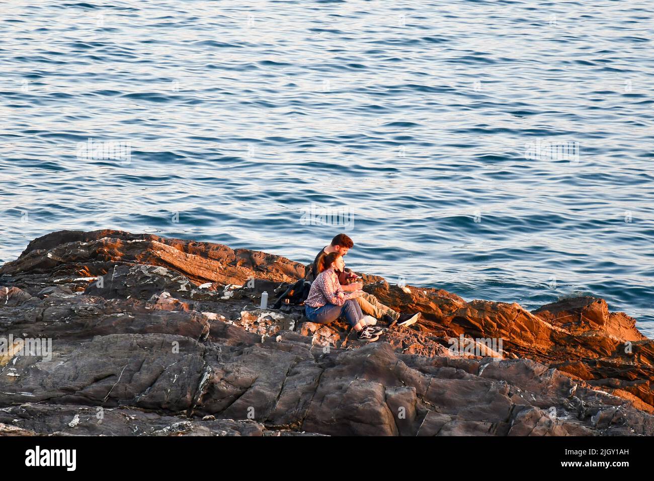 Ein junges Paar (etwa 20 Jahre alt) sitzt auf der Klippe am Meer bei Sonnenuntergang, Nervi, Genua, Ligurien, Italien Stockfoto