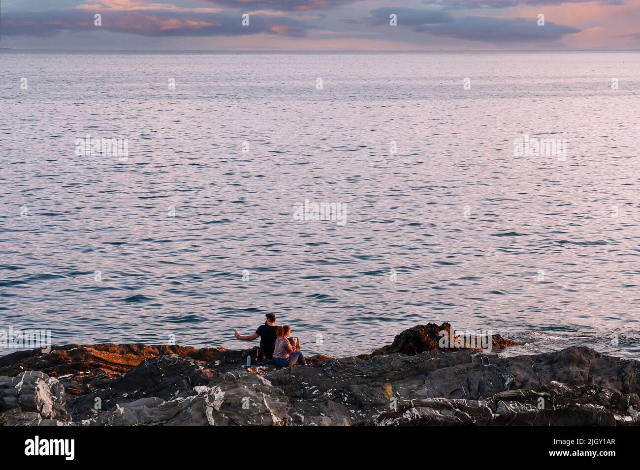 Ein junges Paar (etwa 20 Jahre alt) sitzt auf der Klippe am Meer und macht Selfies mit einem Smartphone bei Sonnenuntergang, Nervi, Genua, Ligurien, Italien Stockfoto