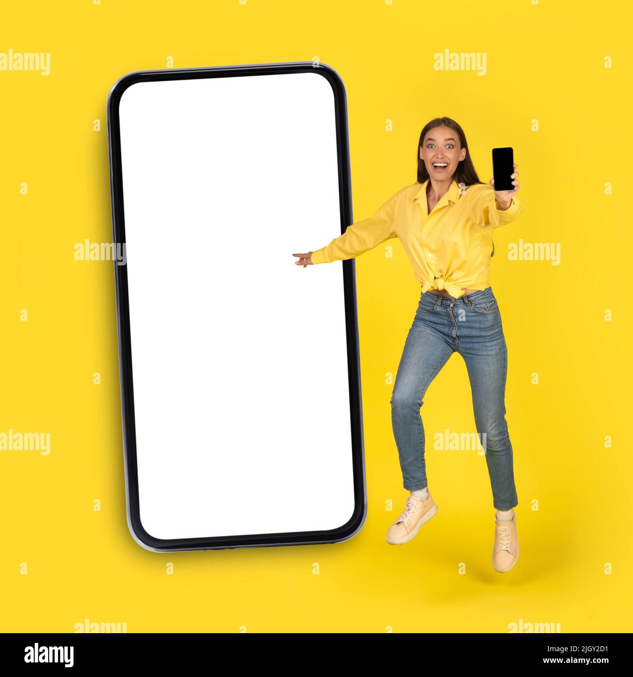 Weiblich Zeigt Smartphone-Bildschirm In Der Nähe Von Riesigen Telefon Über Gelben Hintergrund Stockfoto