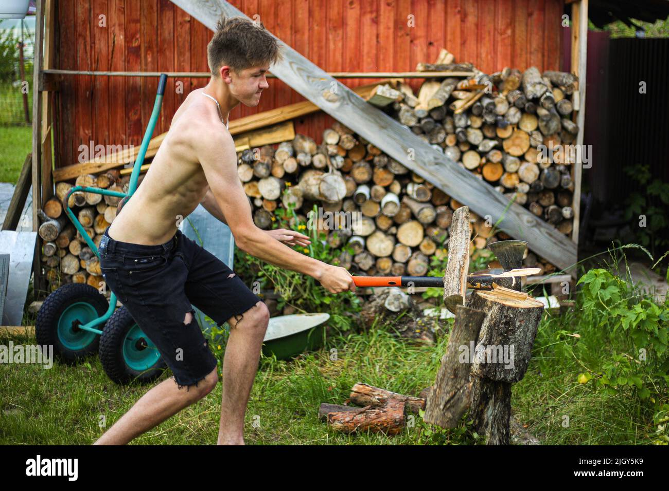 Ein dünner, drahtiger Teenager, barfuß, in schwarzen Shorts, hackt an einem klaren, sonnigen Sommertag mit einer Axt auf einem Stumpf, auf grünem Gras Holz Stockfoto