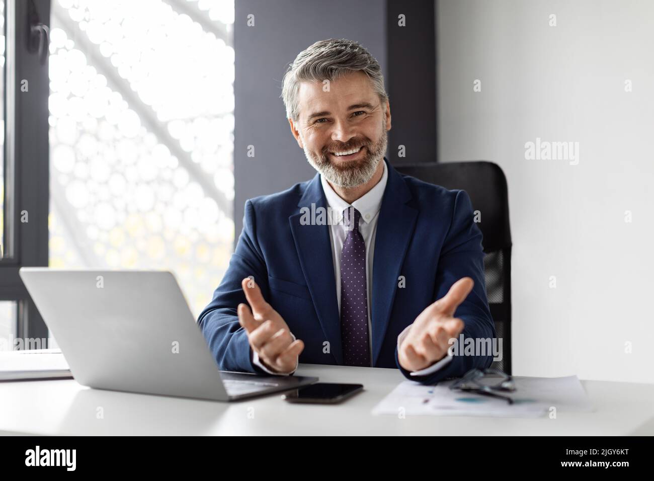 Erfolgreicher Geschäftsmann Mittleren Alters Sitzt Im Büro, Lächelt Und Gestikuliert Vor Der Kamera Stockfoto
