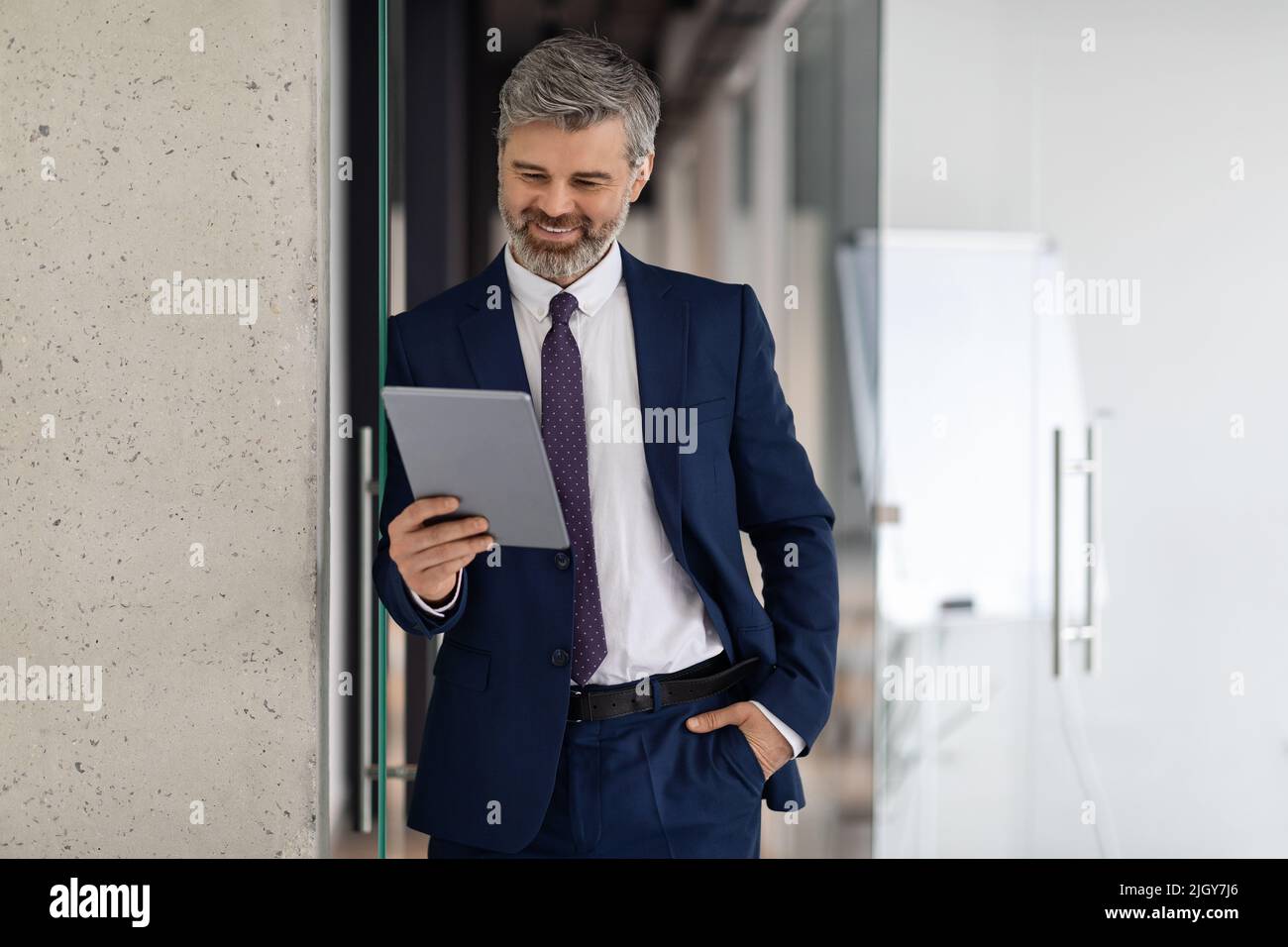 Ein Geschäftsmann mittleren Alters, der ein digitales Tablet benutzt, während er im Korridor des Bürogebäudes steht Stockfoto