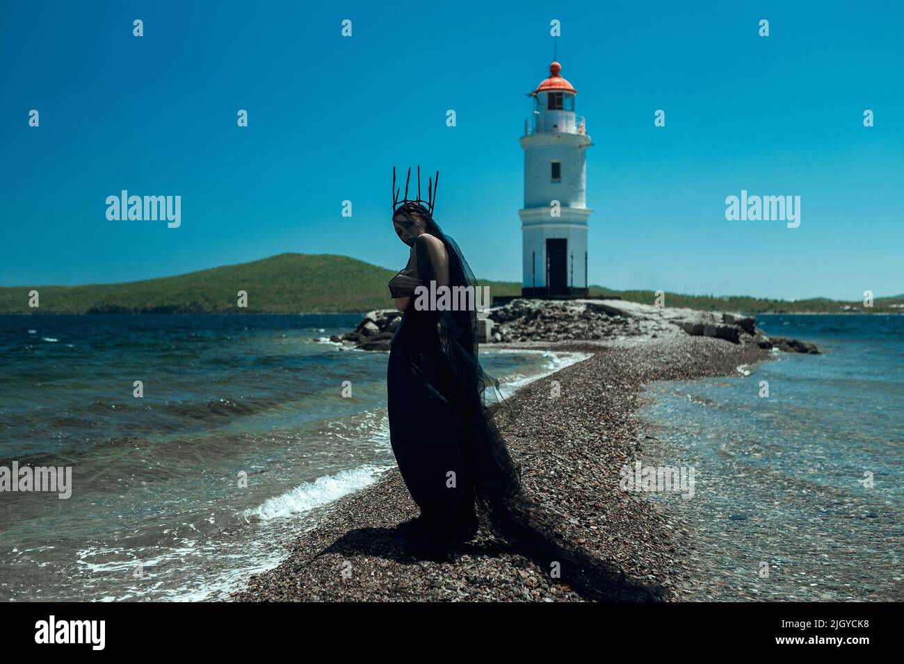 Modell mit schwarzem Gothic-Kleid und dunkler Krone posiert mit einem Leuchtturm Stockfoto