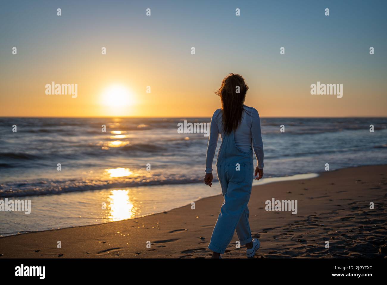 Frau in einem Jeansjumpsuit, die bei Sonnenuntergang am Strand spazieren geht Stockfoto