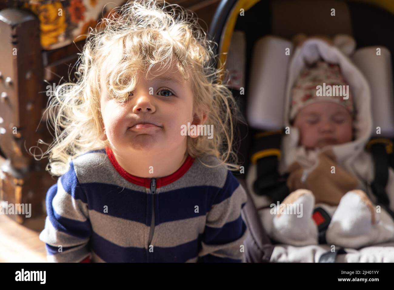 Nahaufnahme mit selektivem Fokus des frechen, zweijährigen kaukasischen Jungen mit welligem blondem Haar und verschwommener neugeborener Schwester im Babysitz im Hintergrund. Stockfoto