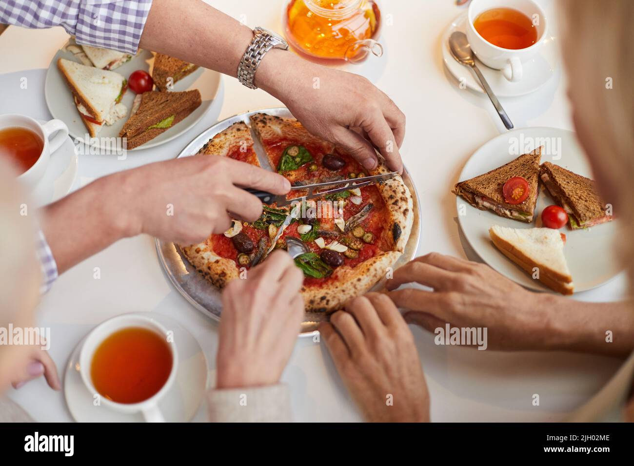 Direkt über der Ansicht eines nicht erkennbaren Mannes mit Armbanduhr, der voll mit Snacks am Tisch sitzt und kleine Pizza für Freunde schneidet Stockfoto
