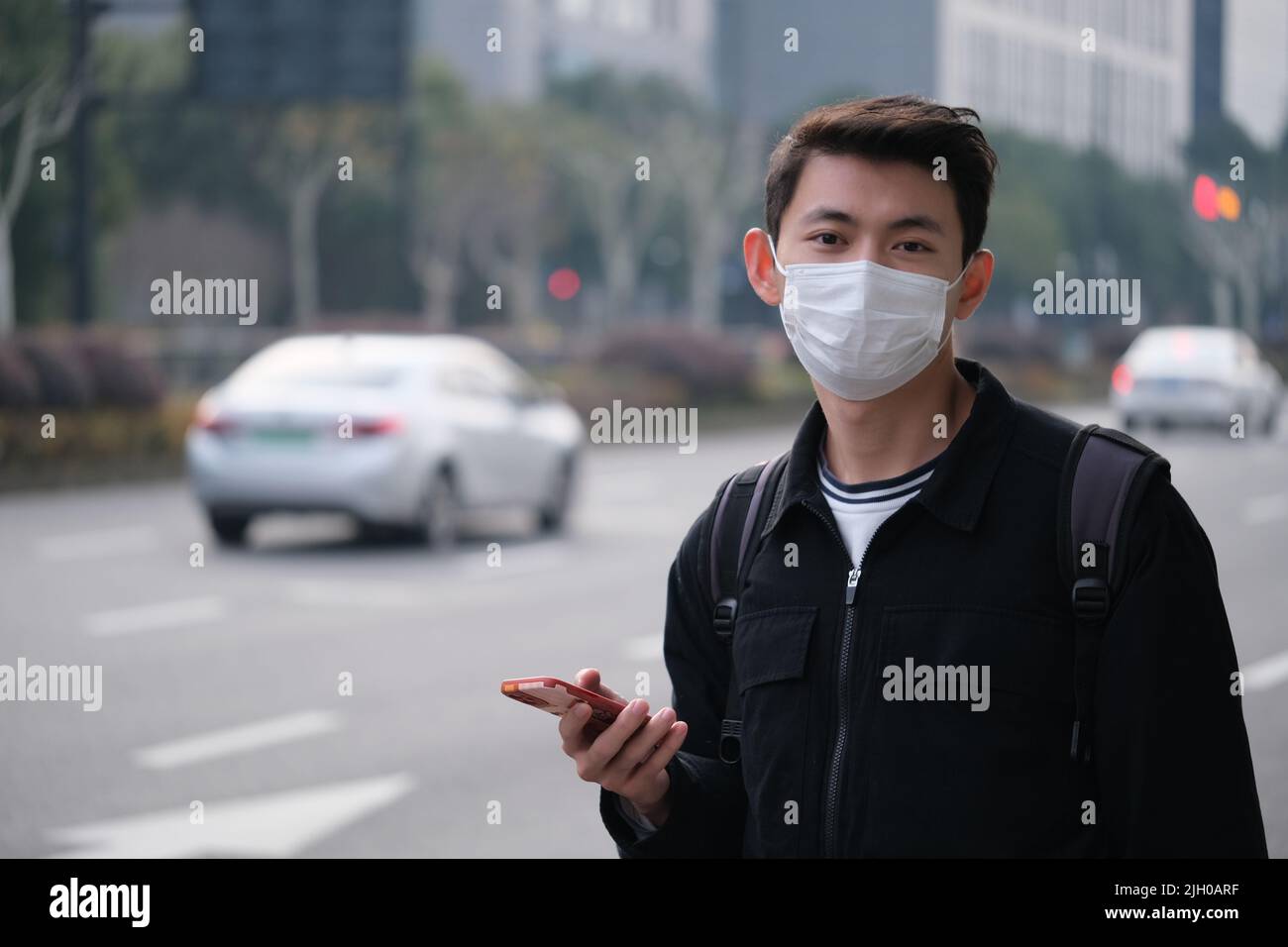 Asiatischer junger Mann mit Gesichtsmaske, der das Telefon hält, um ein Taxi in der Nähe der Straße zu rufen, und in die Kamera schaut Stockfoto