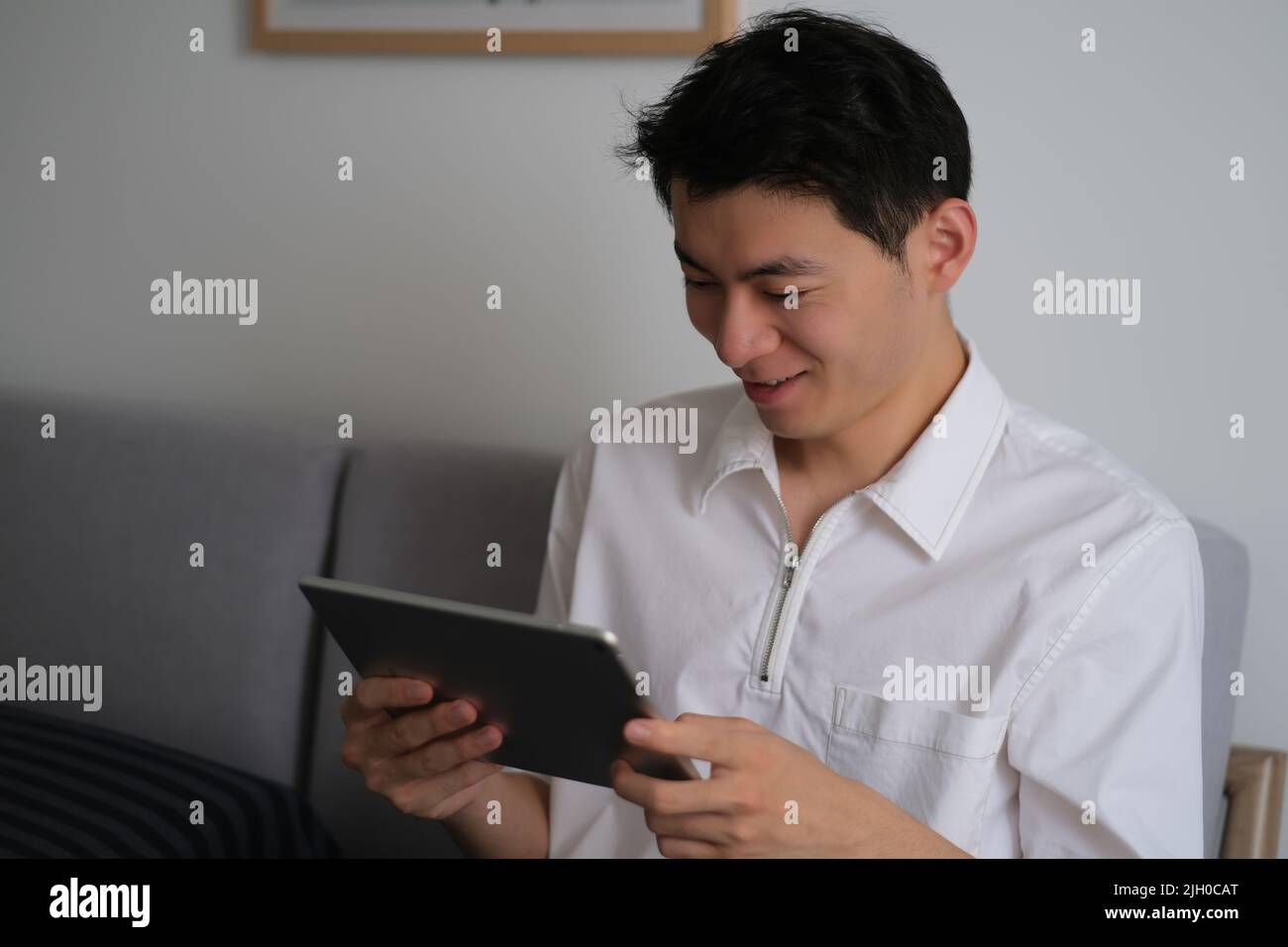 Hübscher junger Mann aus Asien, der zu Hause lächelnd auf einen Tablet-Computer schaut Stockfoto