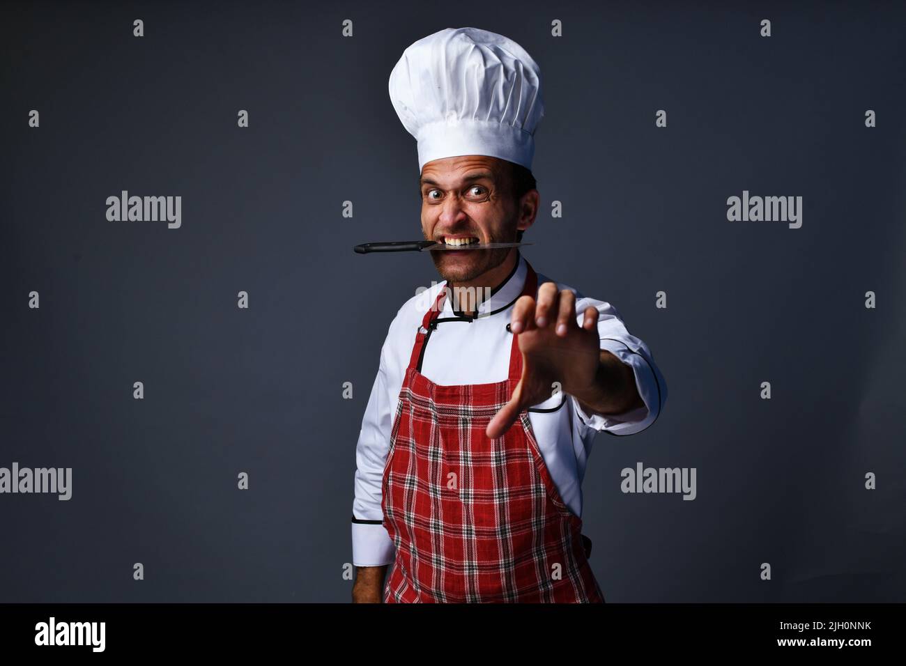 Männlicher Koch posiert wütend Ausdruck mit Messer im Mund Stockfoto