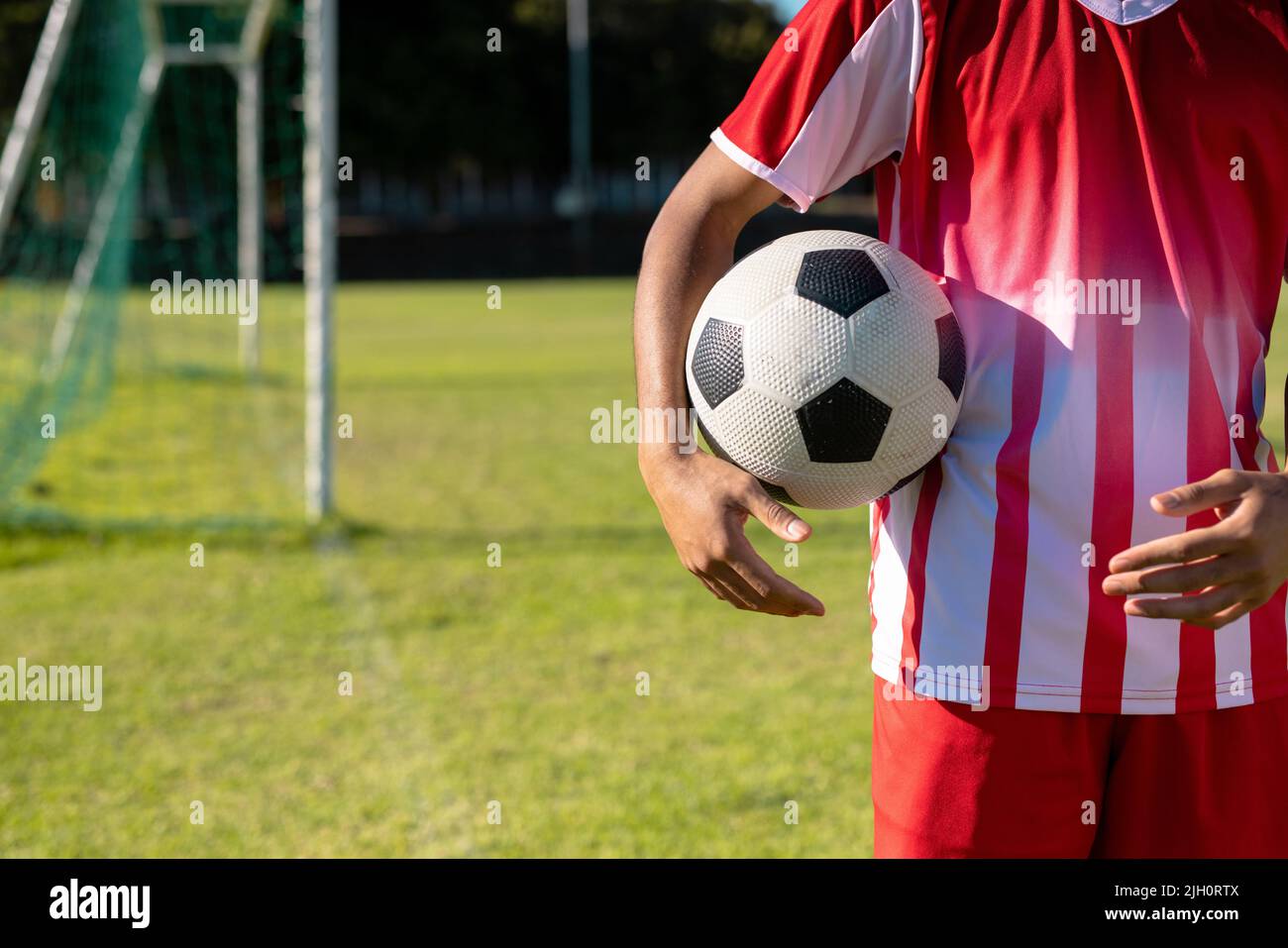 Mittelteil des kaukasischen männlichen Spielers in rotem Trikot mit Fußballball auf dem Spielplatz Stockfoto