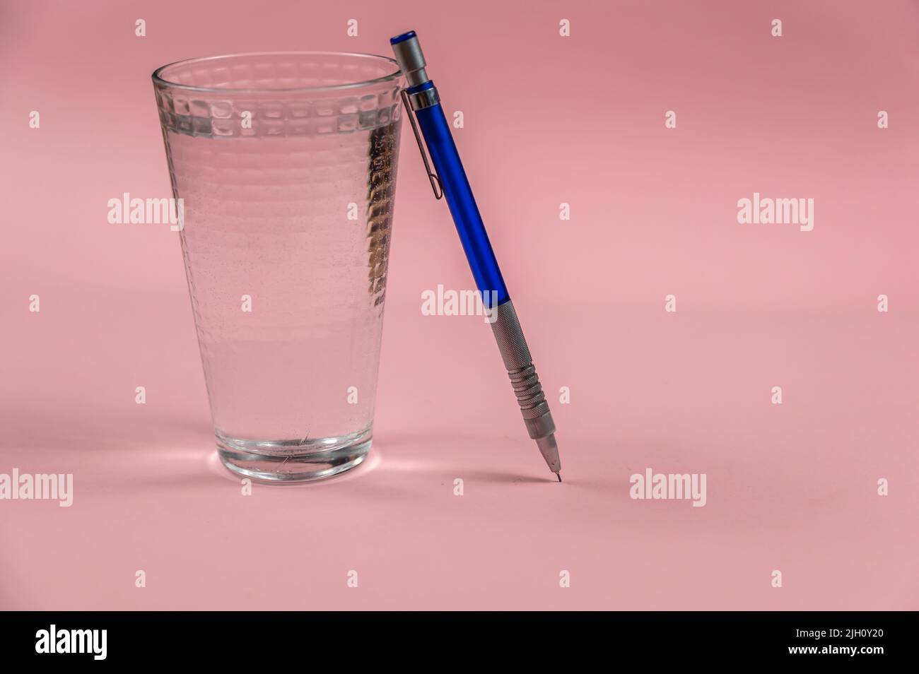 Ein Glas Wasser und ein Bleistift auf einem rosa Hintergrund. Ein volles Glas Trinkwasser. Blauer und silberner Metallic-Bleistift. Kurp-Plan. Selektiver Fokus. Stockfoto