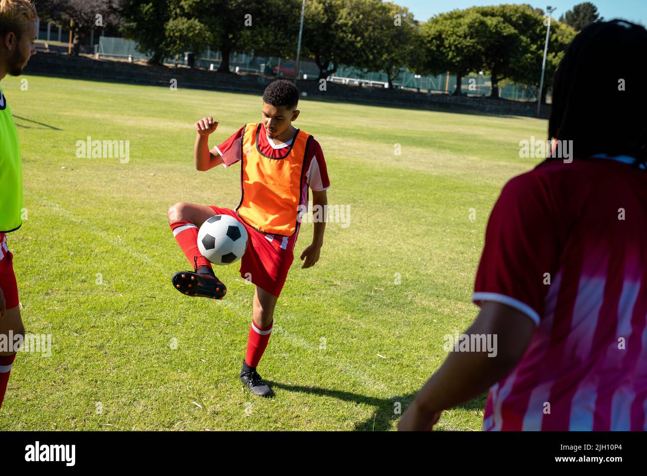Multirassischer junger männlicher Spieler, der Freestyle-Fußball praktiziert, und Teamkollegen, die ihn auf dem Spielplatz angucken Stockfoto