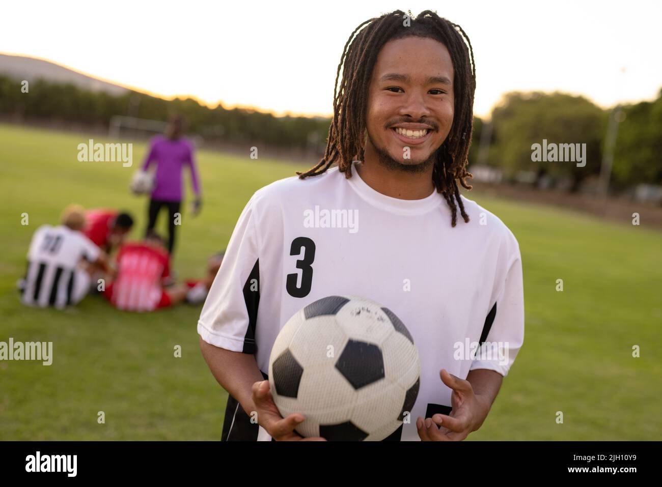 Porträt eines lächelnden männlichen Birazialspielers in weißer Uniform, der den Fußballball auf dem Spielplatz hält Stockfoto