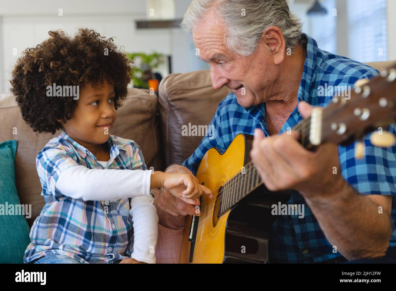 Multirassischer Großvater hält Enkels Hand und lehrt ihn, Gitarre auf dem Sofa zu Hause zu spielen Stockfoto