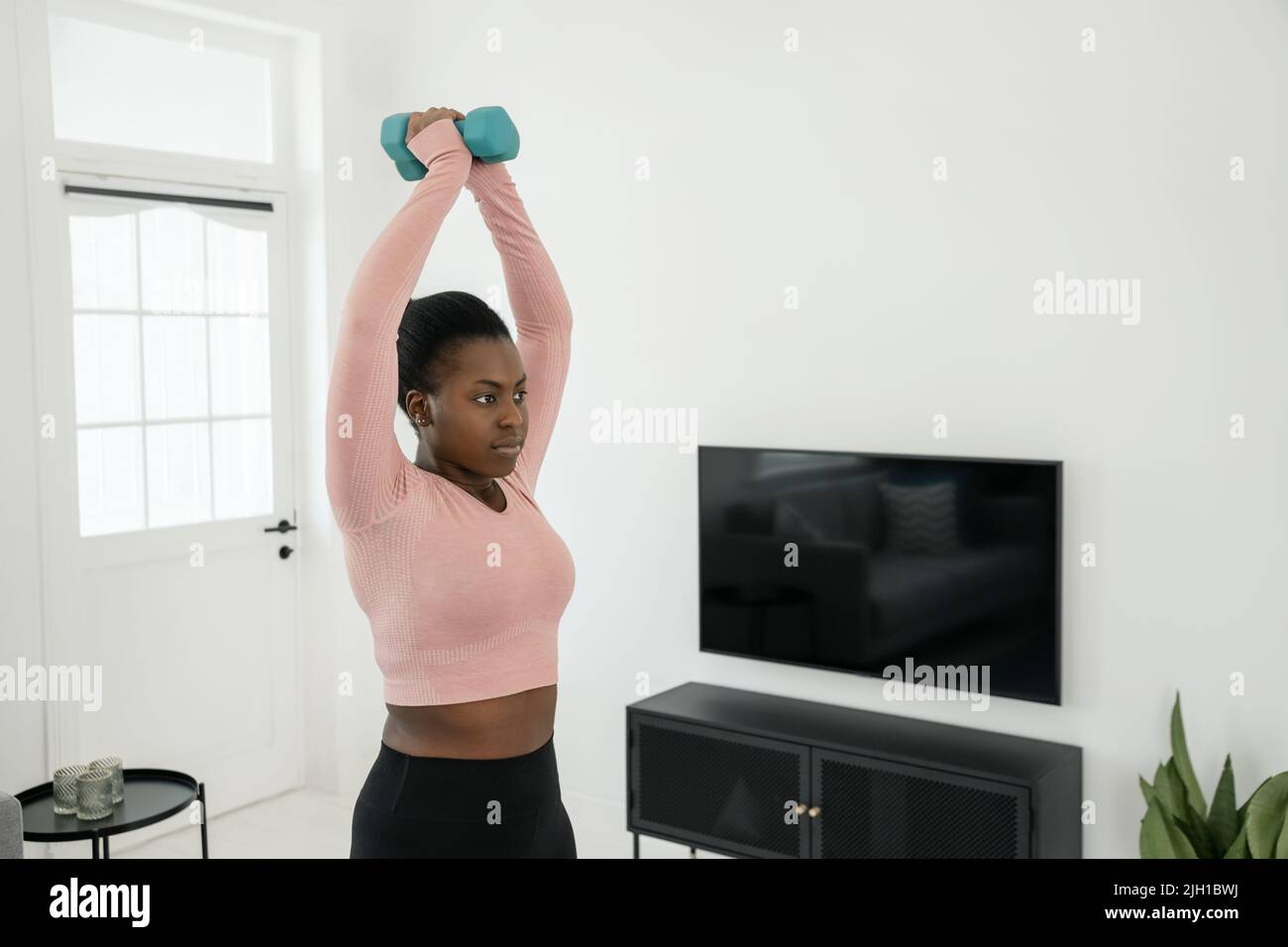 Schwarze afrikanische Frau, die zu Hause trainiert. Beim Hanteltraining mit Gewichten werden die Arme im modernen Wohnzimmer von zu Hause aus angehoben Stockfoto